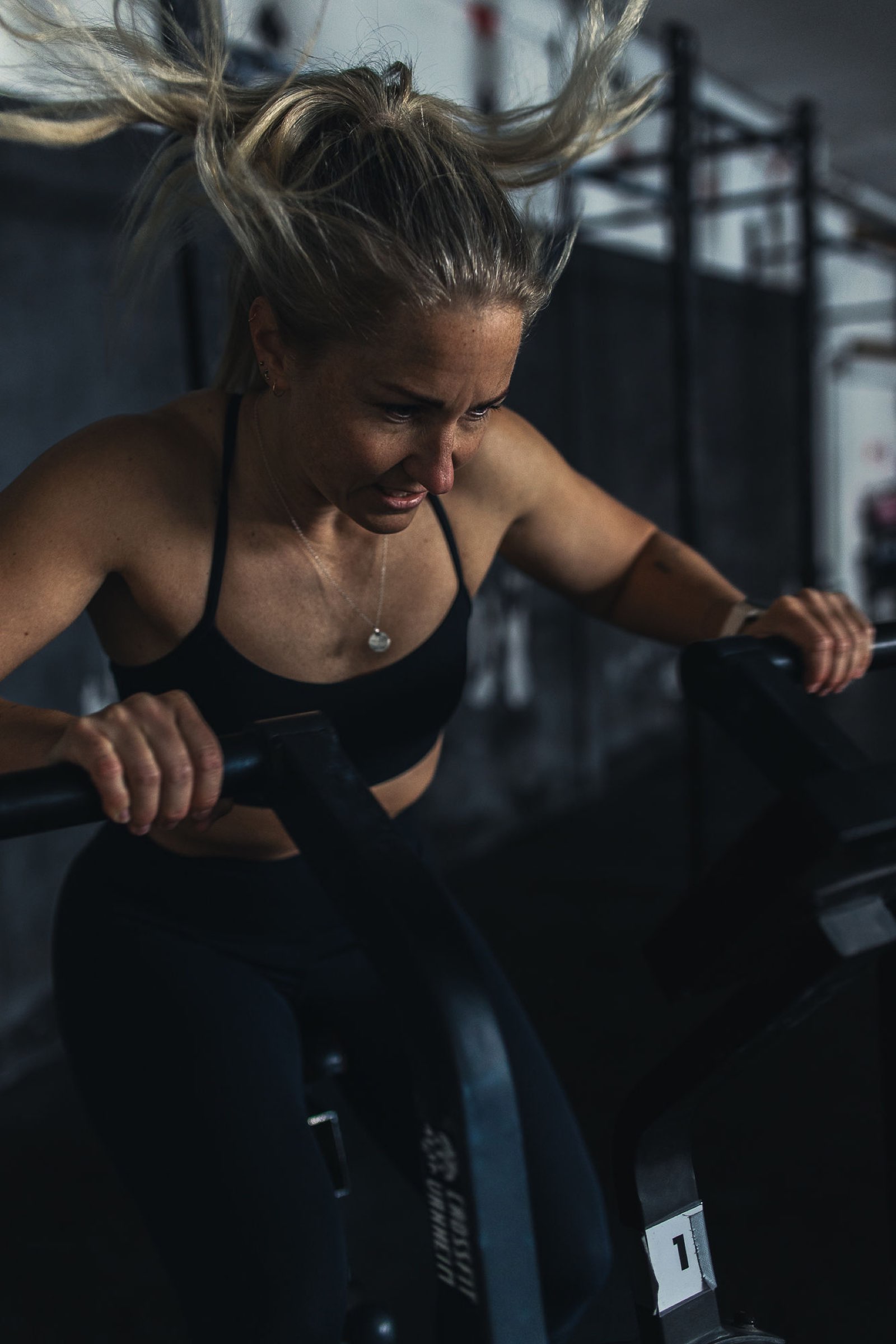 Female athlete at full effort on the air bike during Movement Rebels intervals, hair flying