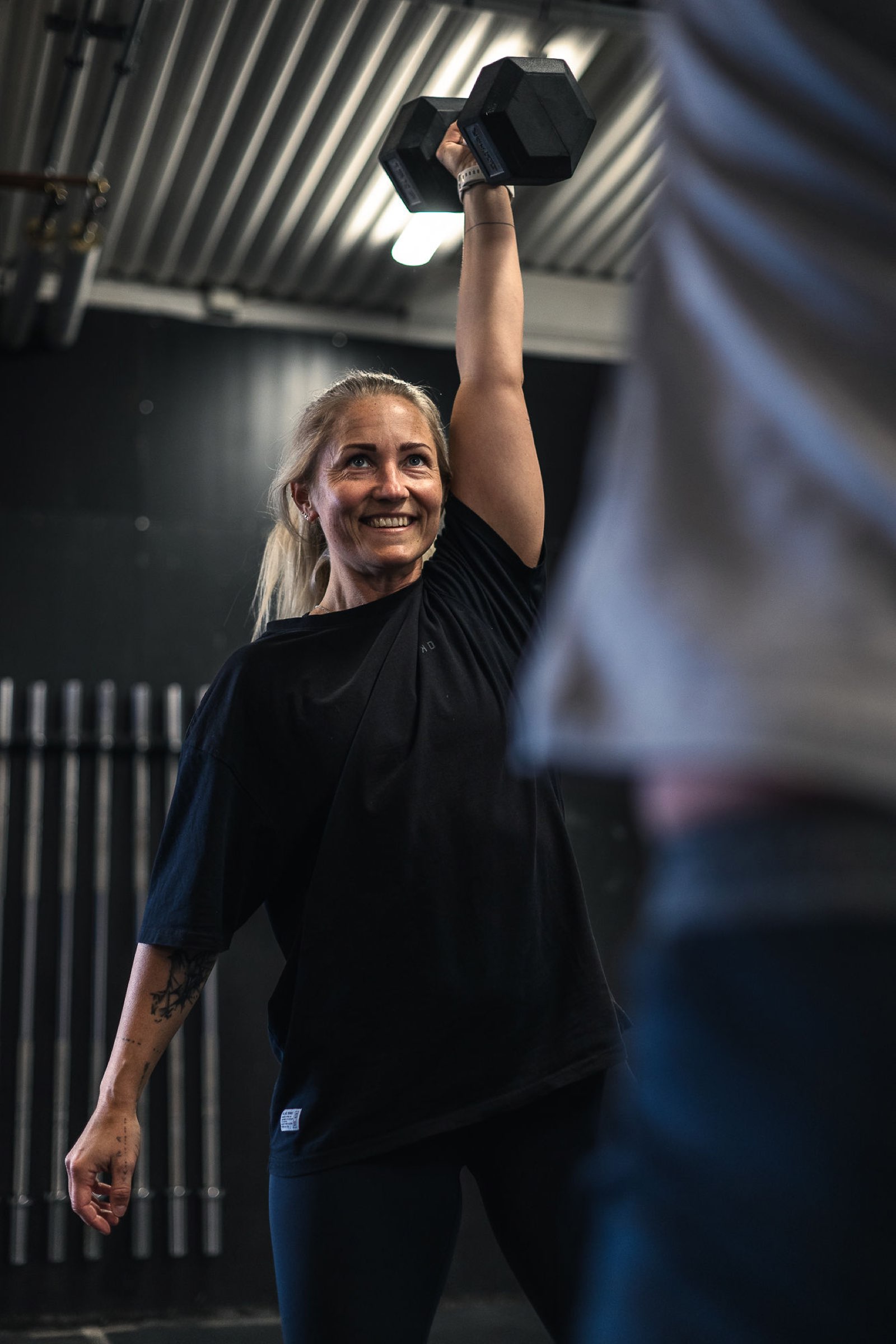 Smiling female athlete locking out a single-arm dumbbell press during a Movement Rebels strength session